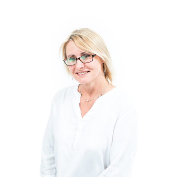 Portret uśmiechniętej blondynki w okularach i białej bluzce, pozującej na jasnym tle.