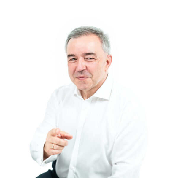 Portret uśmiechniętego mężczyzny z krótkimi siwymi włosami, ubranego w białą koszulę, na jasnym tle.