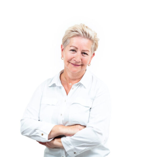 Portret uśmiechniętej kobiety z krótkimi blond włosami, ubranej w białą koszulę, stojącej ze skrzyżowanymi rękami na jasnym tle.