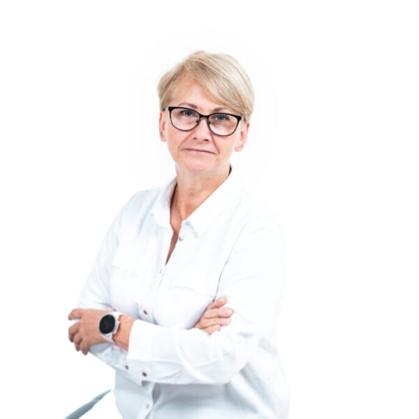 Portret kobiety z krótkimi blond włosami i w okularach, ubranej w białą koszulę, stojącej ze skrzyżowanymi rękami na jasnym tle.
