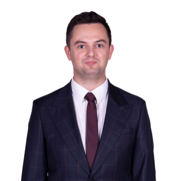 Portret mężczyzny z krótkimi ciemnymi włosami, ubranego w ciemny garnitur, białą koszulę i krawat, na jasnym tle.